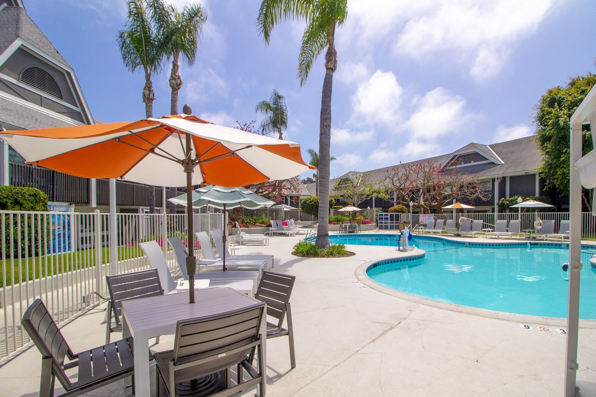 Sunny resort pool with orange-and-white umbrellas, palm trees, and lounge chairs beside a blue swimming pool.