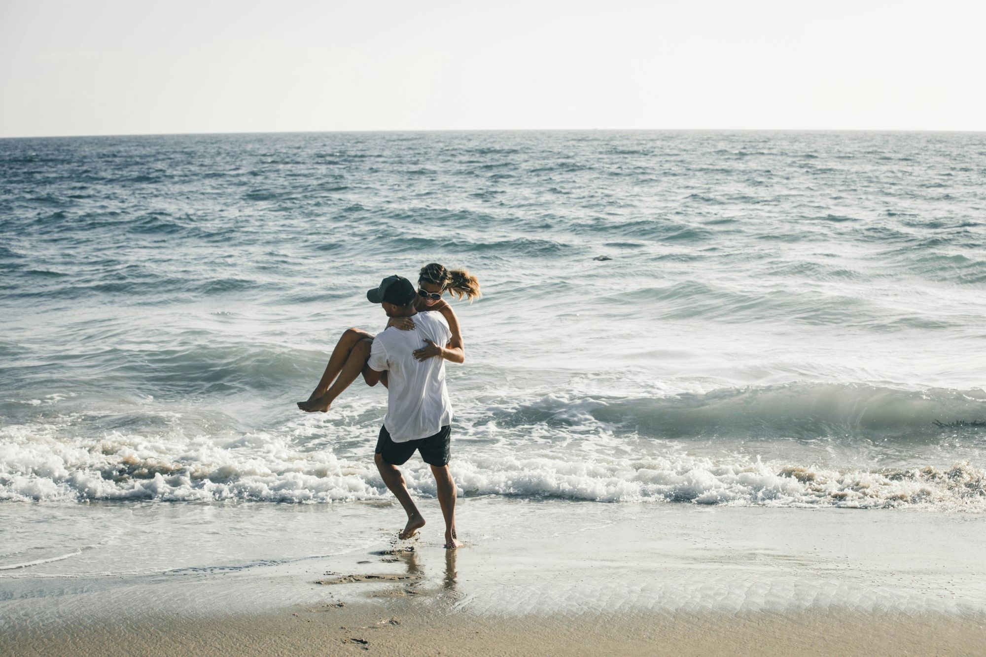 A couple hugs at the shoreline with ocean waves behind them, under a clear bright sky.