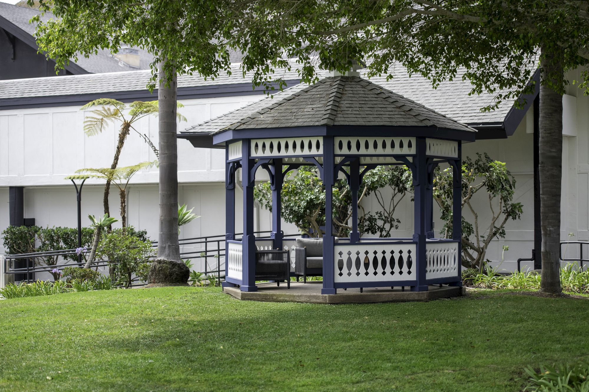 A blue garden gazebo stands on a lawn beside palm trees, under leafy branches in a landscaped yard.