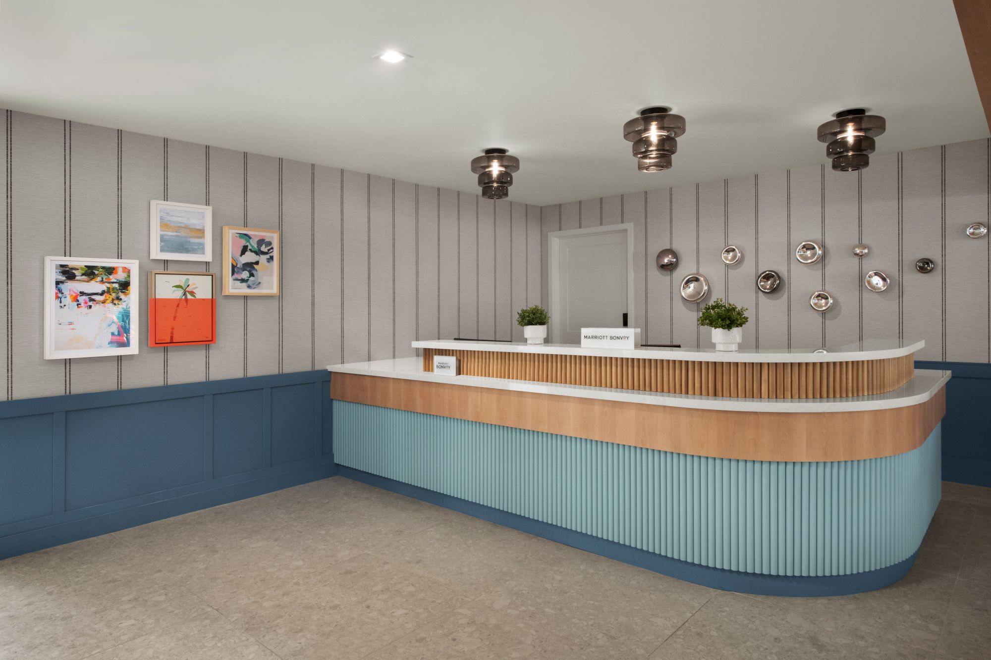 A modern hotel reception area with a curved front desk, framed art, wall sconces, pendant lights, and potted plants.