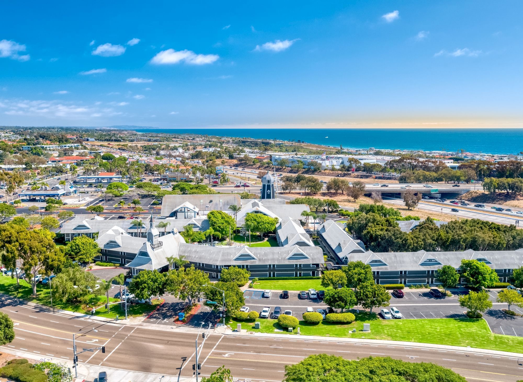 Aerial view of a coastal town: buildings and highways in front, blue ocean on the horizon under a sunny sky.