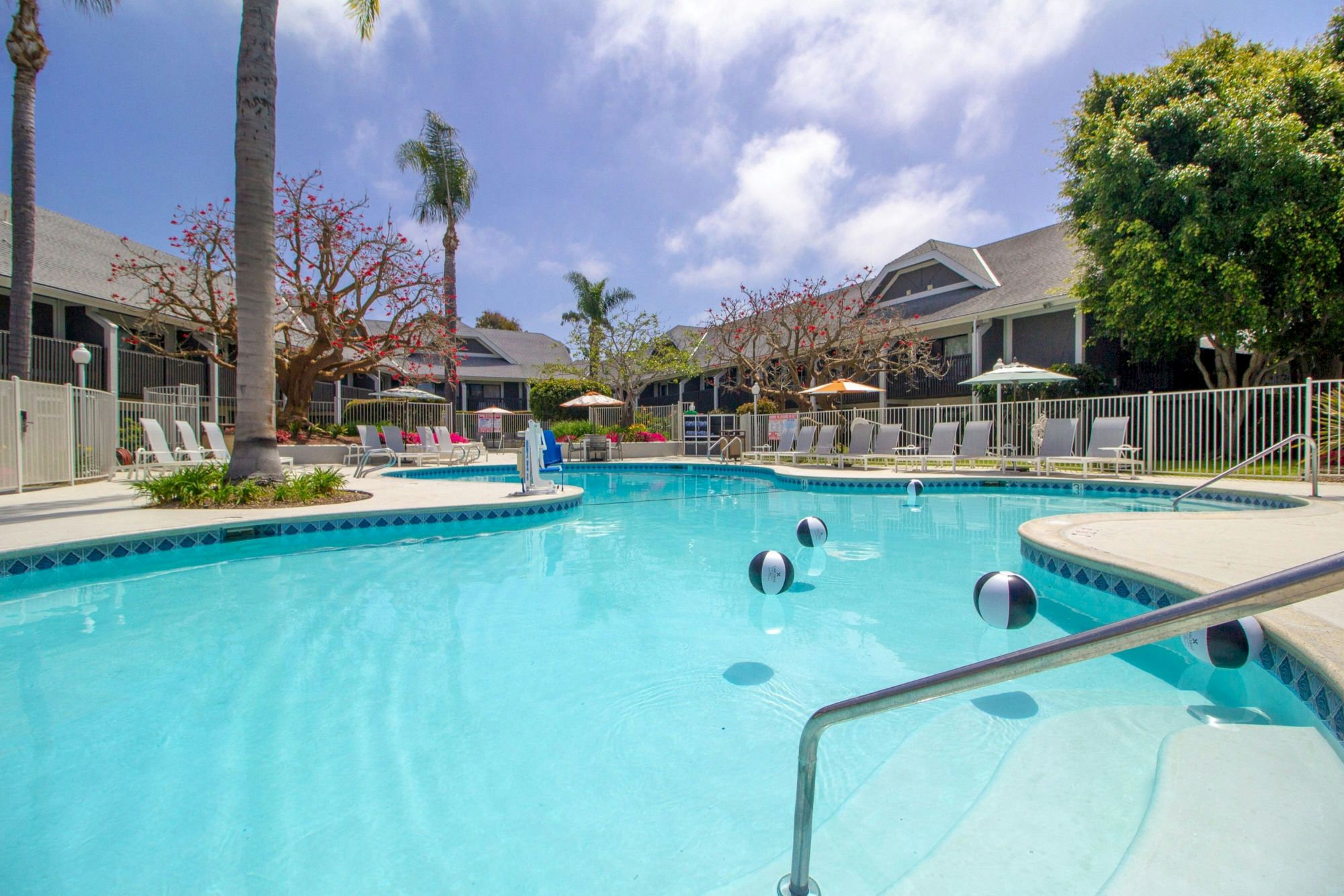 A sunny apartment pool with blue water, lounge chairs, umbrellas, and palm trees by modern buildings.