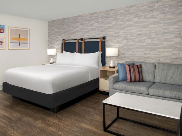 A modern hotel suite with a white bed, dark headboard, gray sofa, lamps, and framed surf-themed wall art.