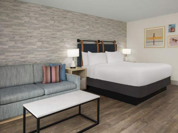 A modern hotel room featuring a king bed with lamps, a gray sofa, a coffee table, and framed beach-themed wall art.