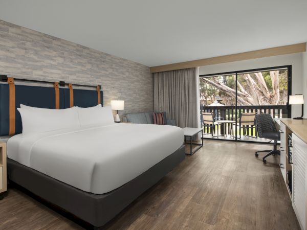 A modern hotel room with a large bed, desk and lamp, and a window/balcony overlooking trees.