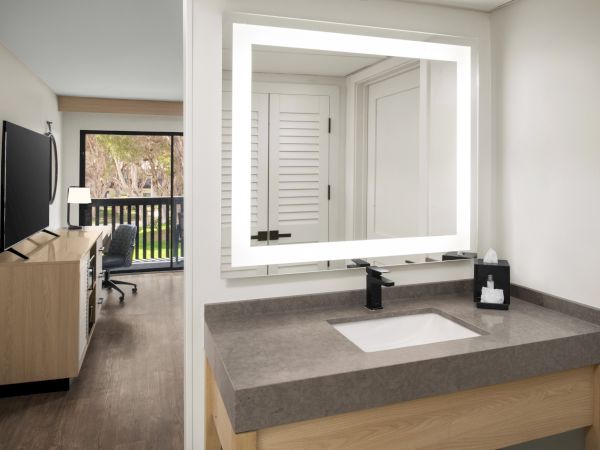 A modern bathroom vanity with a large mirror, sink, and soap, plus a desk area with a TV, chair, and window.