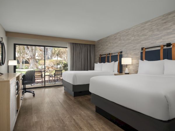 A modern hotel room with two white beds, bedside lamps, and large glass doors overlooking trees outside.