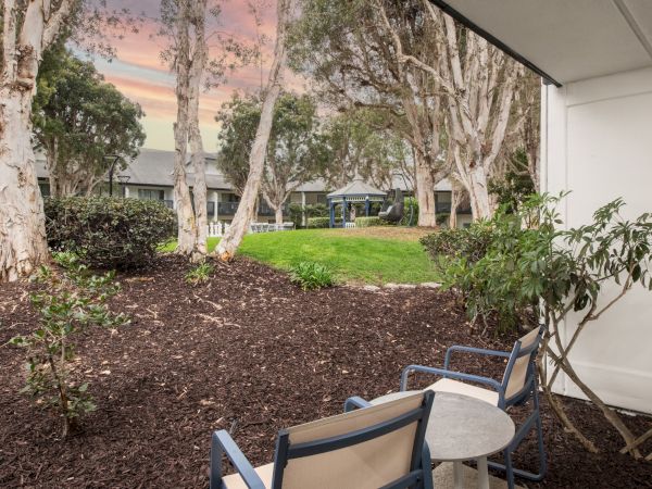 A quiet courtyard patio with birch-like trees, green lawn, outdoor chairs, and a small gazebo beside a walkway at dusk.