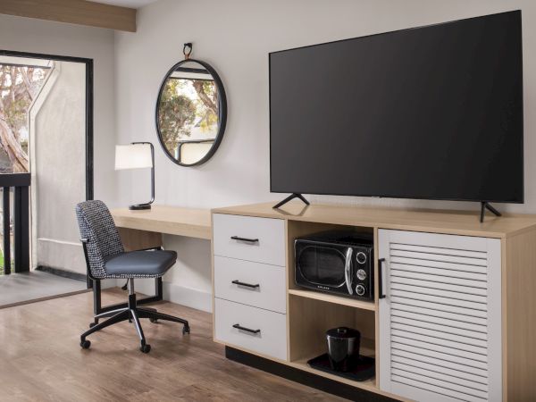 A modern room setup with a flat-screen TV on a console, a desk chair, a wall mirror, and a microwave.