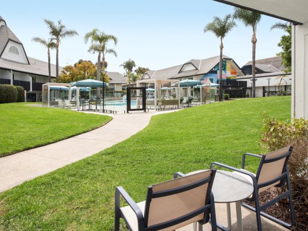 A sunny apartment courtyard with a fenced pool, lounge chairs, umbrellas, lush grass, and tall palm trees beside colorful buildings.