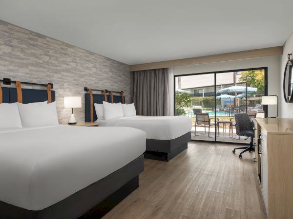 Modern hotel room with two beds, a desk and TV, opening to a patio with chairs, umbrellas, and a poolside view.