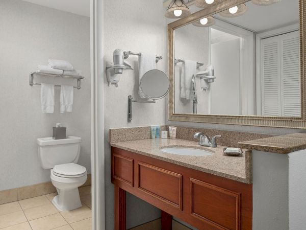 A bright hotel bathroom with a vanity sink, large mirror, towels on racks, wall-mounted fixtures, and a toilet in view.