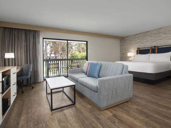 A modern hotel room with a king bed, gray sofa, coffee table, desk, lamps, and floor-to-ceiling windows opening to a balcony.