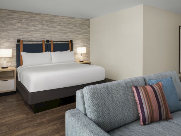 A modern hotel bedroom with a large bed, bedside lamps, stone-pattern wall, and a sofa in the foreground.