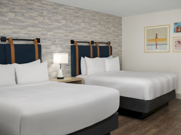 A modern hotel room with two white beds, blue-and-brown headboards, a lamp, and framed wall art.