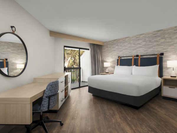 Modern hotel room with a round mirror, wooden desk, bed, nightstands, and a window with curtains.