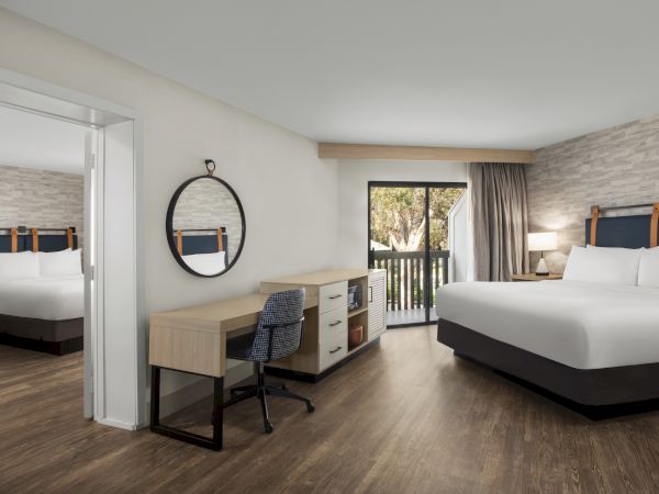 Modern hotel room with two beds, a desk, round wall mirror, and curtains framing a balcony door.