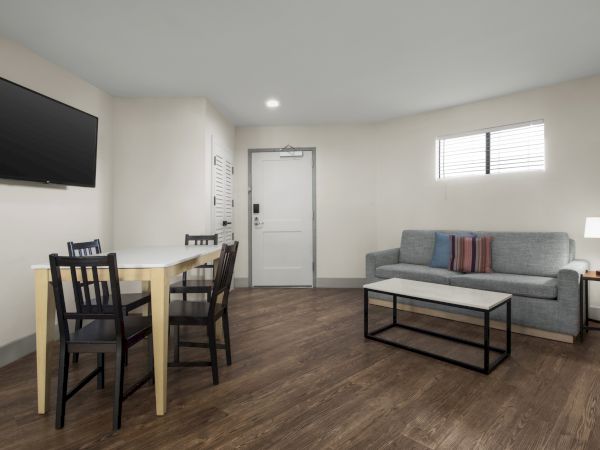 A bright living room with a gray sofa, coffee table, dining set, wall TV, and a white door and blinds.