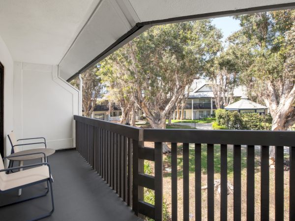 A quiet apartment balcony with two chairs, looking out over a garden of tall trees and nearby buildings.