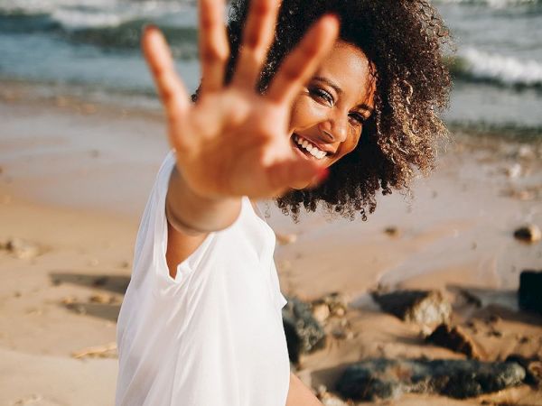 A smiling woman sits on a sunny beach by the ocean, waving her hand toward the camera with waves in the background.
