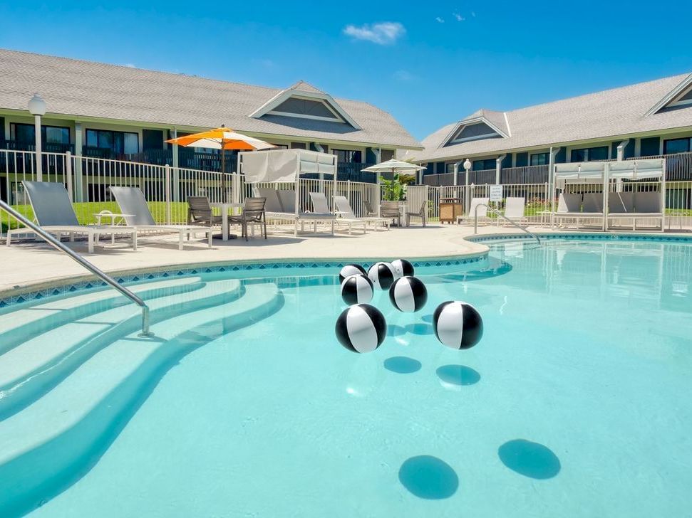 An outdoor motel pool with lounge chairs, umbrellas, and a black-and-silver floating object in the clear blue water.