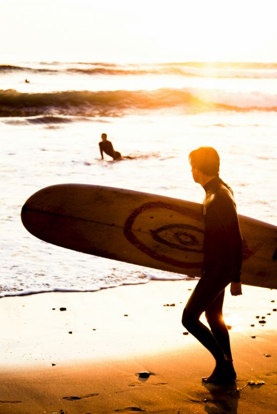 A surfer walks along the beach with a surfboard at sunset, waves rolling in behind them.
