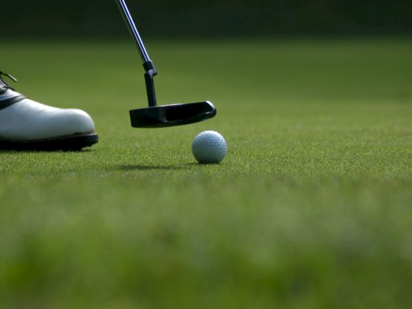 A golfer&rsquo;s shoe and club hover over a golf ball on the green, ready for a putt.