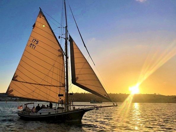 A sailboat with two large sails cruises on a river at sunset, with sun rays reflecting on the water.