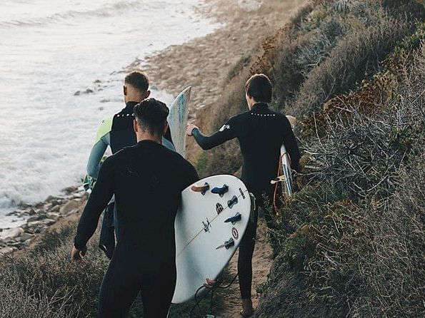 A group of surfers in wetsuits walks along a rocky coastline carrying surfboards near the shore.