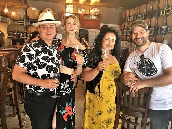 Four people in a winery tasting room, holding wine glasses and smiling, with barrels and warm lights in the background.