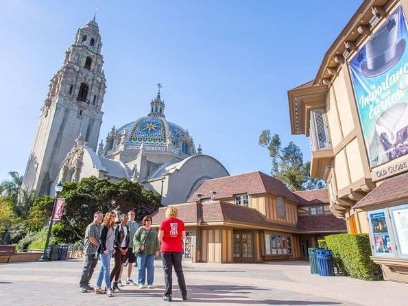 A group of tourists walk in a sunny plaza beside a large dome-and-tower church and a theater marquee, under a clear blue sky.