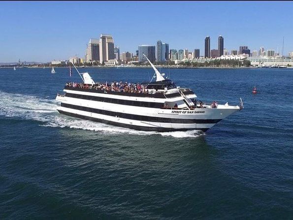 A passenger ferry, the Spirit of San Diego, cruises on the water toward a skyline under a clear blue sky.