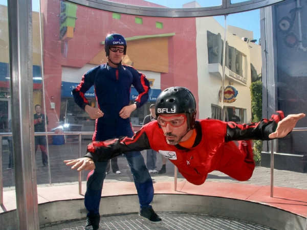 A helmeted man in a red wingsuit is launching off a trampoline while another person stands behind him in an indoor wind setup.