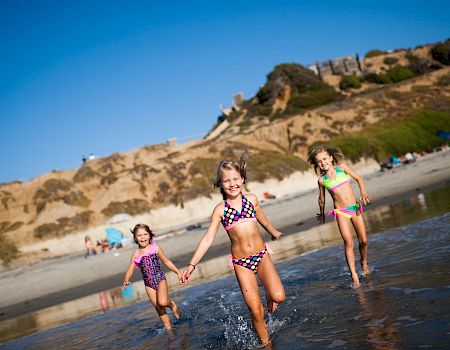 Three kids in colorful swimsuits run through shallow waves on a sunny beach with cliffs and spectators behind them.