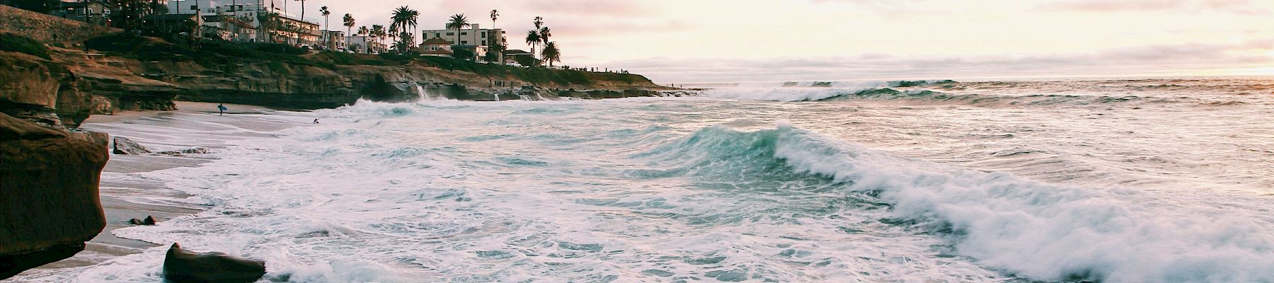 A coastal sunset with crashing waves, rocky shore, and palm-lined homes in the distance.
