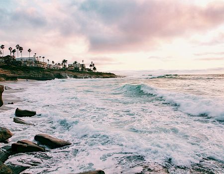 A coastal sunset with crashing waves, rocky shore, and palm-lined homes in the distance.