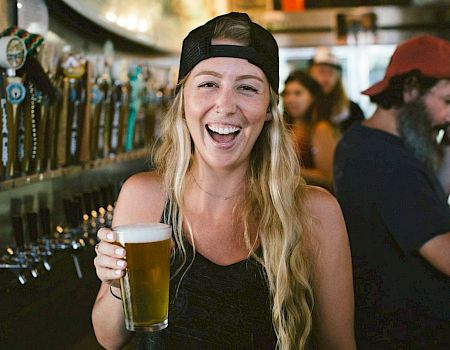 Smiling woman in a bar wearing a backward cap holds a glass of beer while friends chat nearby.