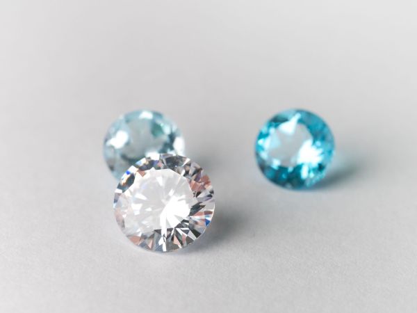 Two gemstones&mdash;a blue round stone and a clear oval diamond-like gem&mdash;rest on a light background, catching bright sparkle reflections.