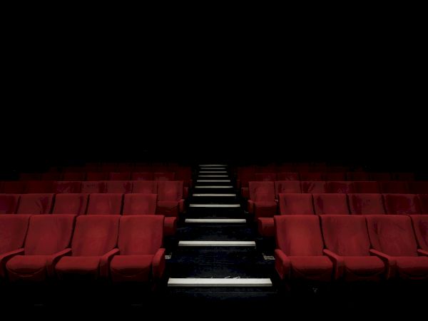 An empty cinema auditorium with rows of red cushioned seats and a central aisle leading to the dark stage.