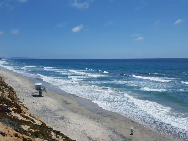 A wide sandy coastline with crashing waves, a blue ocean, and a lifeguard stand under a clear blue sky.