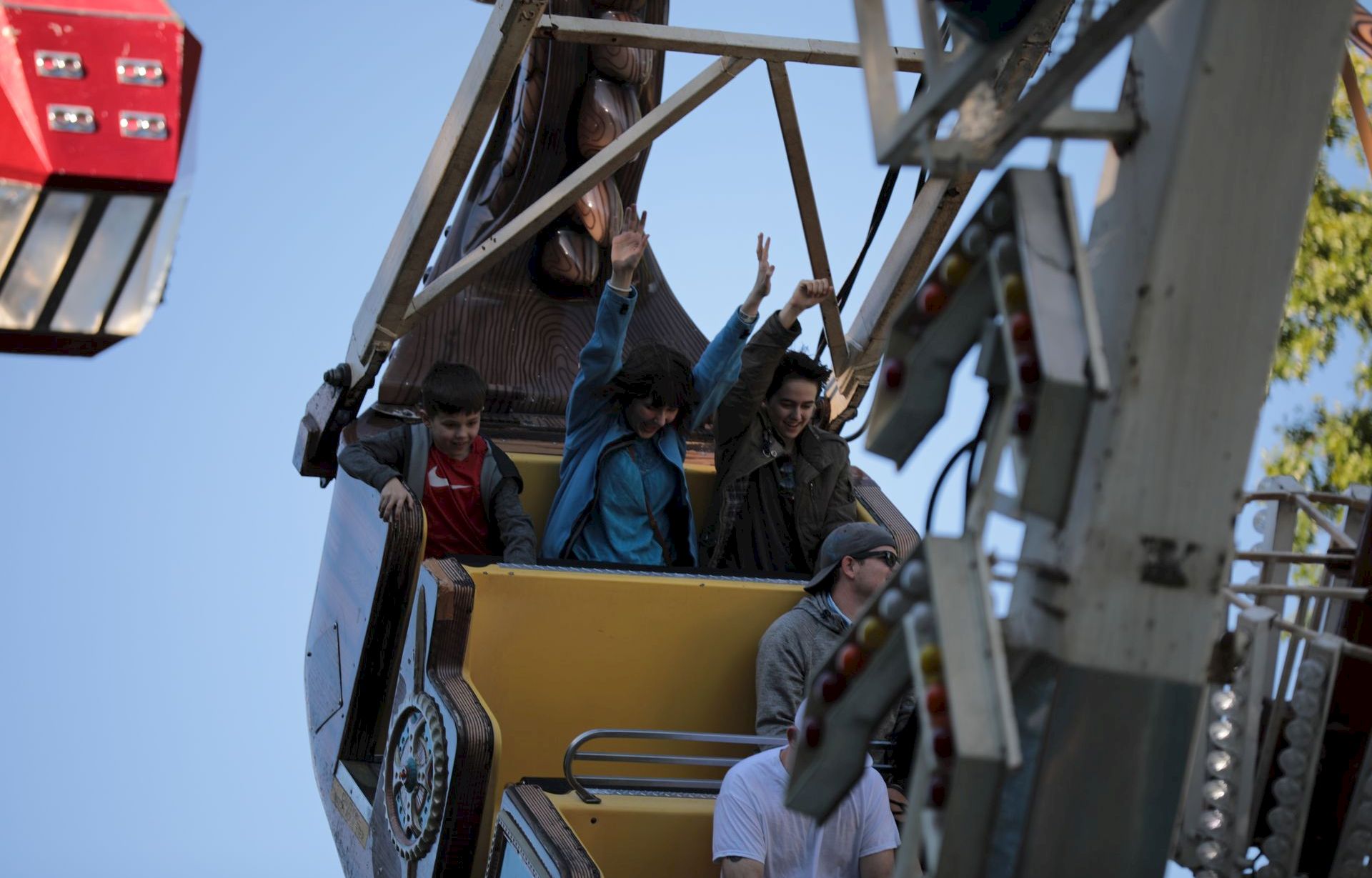 Kids excitedly wave from the platform of a big outdoor ride as a worker stands nearby, taking in the view.