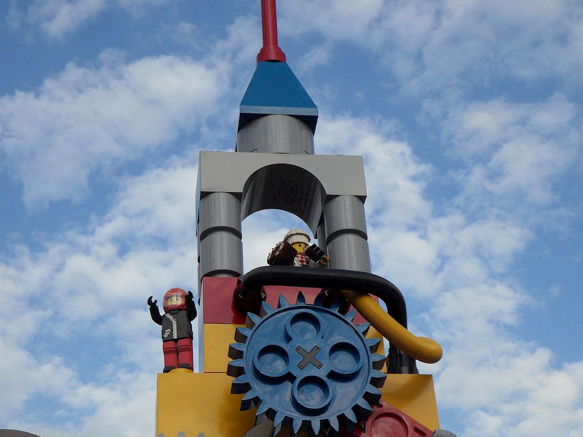 A LEGO-style robot with gears and a waving green/red flag stands against a bright cloudy sky.