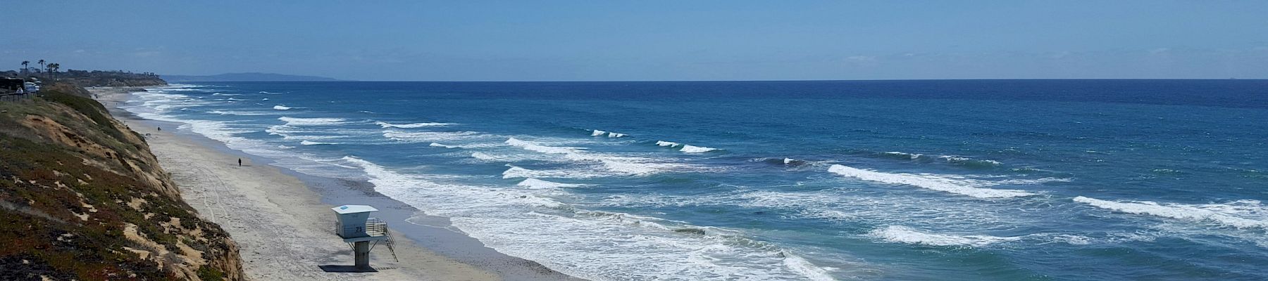 A scenic coastline beach with crashing waves, a lifeguard stand, sandy shore, and a bright blue sky.