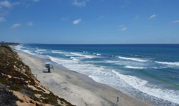 A scenic coastline beach with crashing waves, a lifeguard stand, sandy shore, and a bright blue sky.