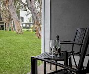 A patio table with two wine glasses and a bottle of sparkling wine sits beside a grassy yard and trees.