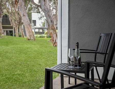 A patio table with two wine glasses and a bottle of sparkling wine sits beside a grassy yard and trees.