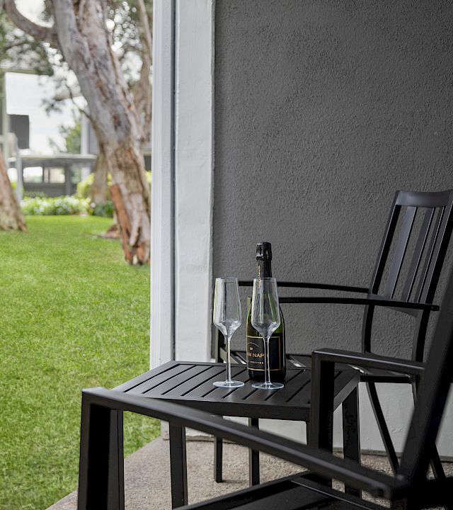 A patio table with two wine glasses and a bottle of sparkling wine sits beside a grassy yard and trees.