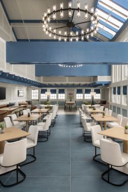 A bright modern lounge with rows of tables and white chairs beneath a large chandelier and skylights.