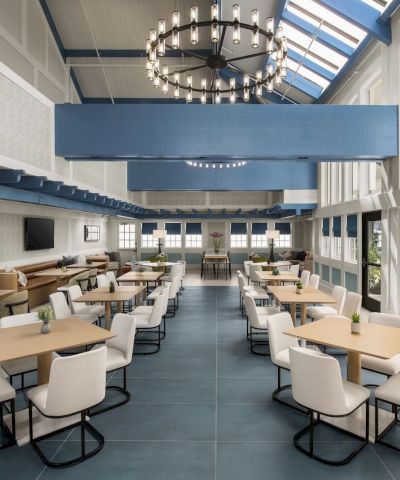 A bright modern lounge with rows of tables and white chairs beneath a large chandelier and skylights.