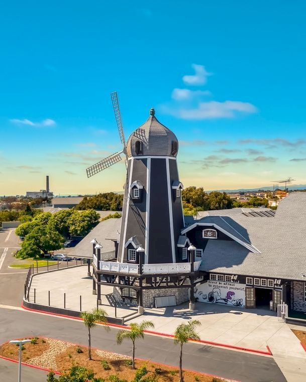 A tall Dutch-style windmill stands on a hill beside buildings under a bright blue sky.